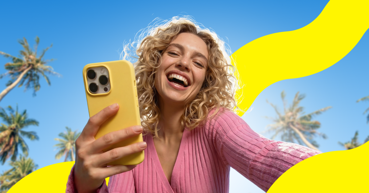 A smiling traveller holding up her phone under palm trees