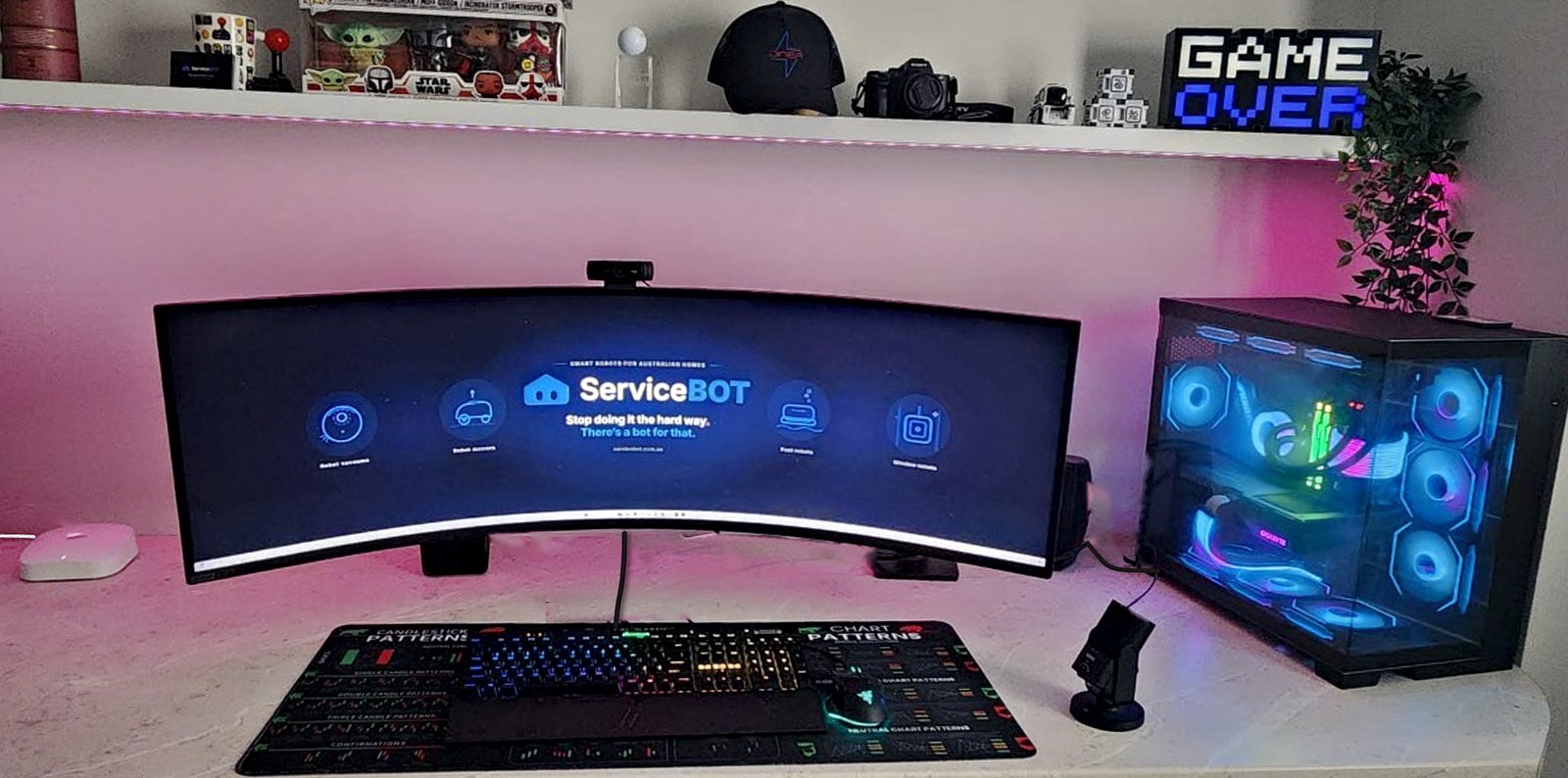 ServiceBot founder Ed's home office in Sydney: a curved ultrawide monitor showing the ServiceBot logo, a gaming PC and collectibles on the shelf
